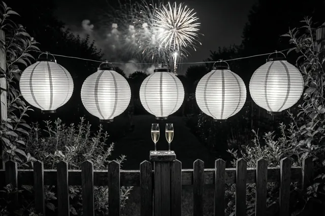 Op een zwart-witfoto hangen vijf brandende papieren lampionnen boven een houten schutting, met twee champagneglazen op een paal en vuurwerk aan de nachtelijke hemel.