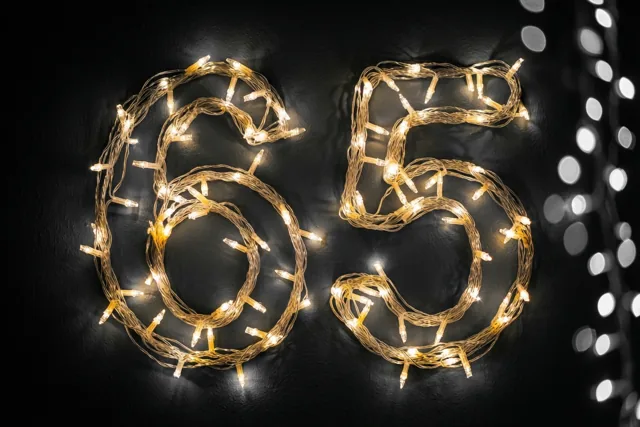 Het getal 65 is gevormd met warme lichtjes tegen een donkere muur, met wazige lichtpuntjes op de achtergrond.