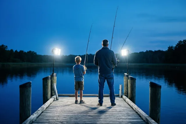 Een vader en zoon staan van achteren gezien bij schemering te vissen op een houten steiger die fel wordt verlicht, tegen een diepblauwe lucht en water.
