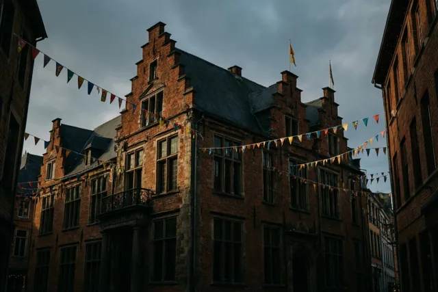Kleurrijke carnavalsvlaggen hangen over de straat voor een historisch bakstenen stadhuis met trapgevels onder een bewolkte hemel.