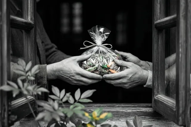 Twee paar handen geven een voedselpakket in doorzichtig plastic door via een open houten raam, op een grotendeels zwart-witte foto met kleur in het pakket en het groen op de voorgrond.