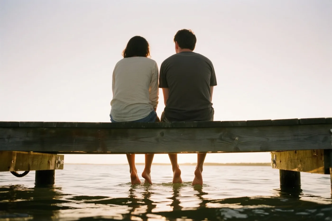 Twee mensen zitten met hun rug naar de kijker op een houten steiger met hun blote voeten in het water in warm goudkleurig licht.