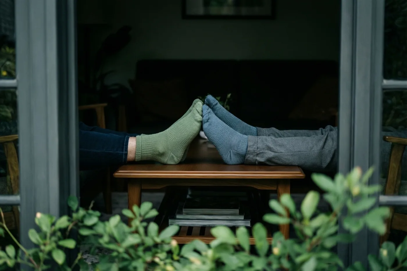 Twee paar voeten in niet bij elkaar passende sokken liggen naast elkaar op een houten salontafel, gezien door een raam met groen op de voorgrond.