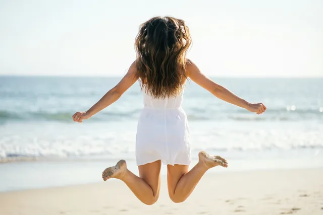 Een persoon met lang donker haar en een witte jurk springt op een zandstrand, van achteren gezien met zand aan de blote voeten en de zee op de achtergrond.