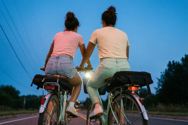 Twee vrienden zijn van achteren te zien tijdens een fietstocht bij blauwe schemering, waarbij één op de bagagedrager van de ander zit en fietslichten en kleding warme accenten geven.