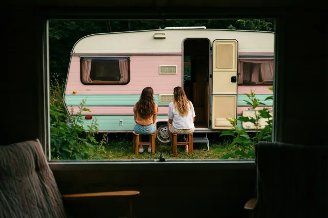 Door een raam zijn twee vrienden op houten krukjes te zien bij een oude caravan met verbleekte roze en mintgroene strepen, omringd door groen.