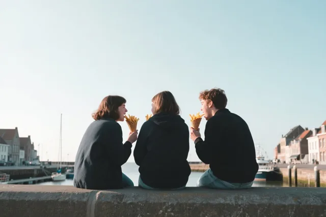 Drie vrienden zitten met hun rug naar de kijker op een havenmuur in een kuststadje, elk met een papieren puntzak frieten in de hand.