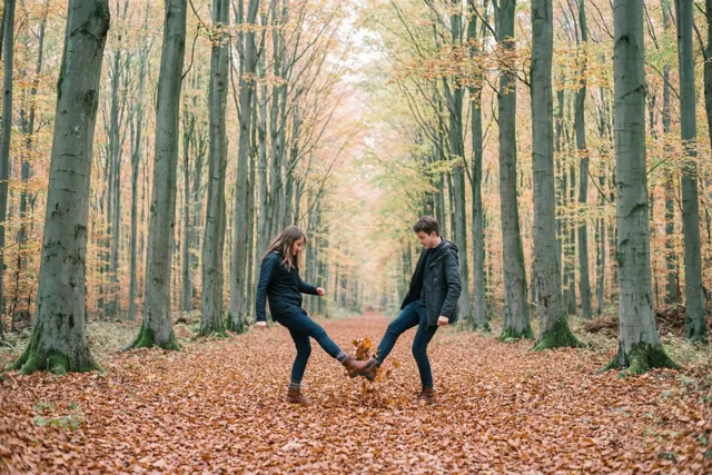 Twee vrienden schoppen speels tegen herfstbladeren op een pad tussen hoge beuken in warm herfstlicht.