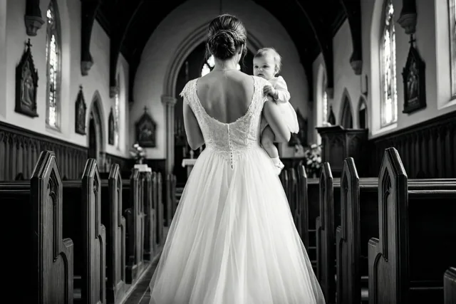 Een zwart-witfoto toont een vrouw in een witte trouwjurk van achteren terwijl ze een baby in een witte doopjurk door het gangpad van een kerk draagt.