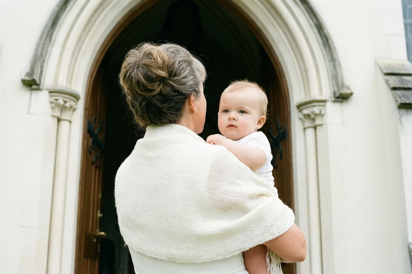 Een vrouw met grijs haar staat met haar rug naar de camera bij een houten kerkdeur met boog en houdt een baby in een witte omslagdoek vast.