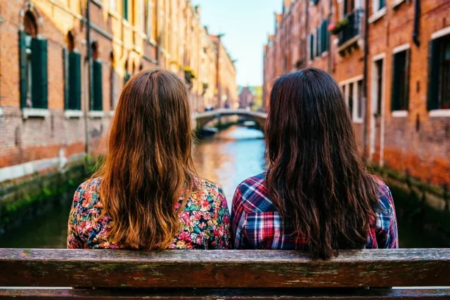 Twee vrouwen met lang haar zitten met hun rug naar de kijker op een houten bank aan een kanaal tussen oude bakstenen gebouwen in warm, goudkleurig licht.