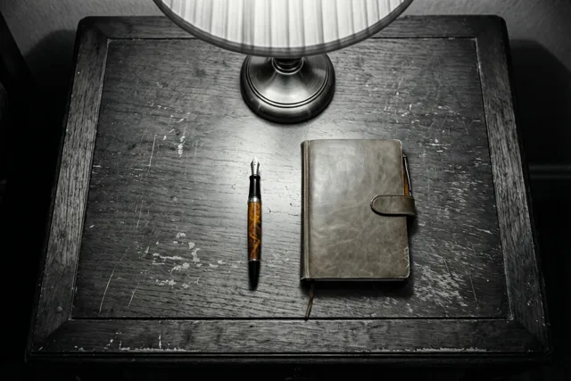 Een vulpen en een notitieboek met leren kaft liggen op een versleten houten bureau onder een lamp met een geribbelde kap.