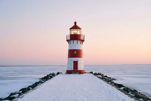 Een kleine rood-witte vuurtoren met warm brandend licht staat aan het einde van een besneeuwde pier bij een bevroren zee, onder een zachtroze en bleekblauwe schemerhemel.