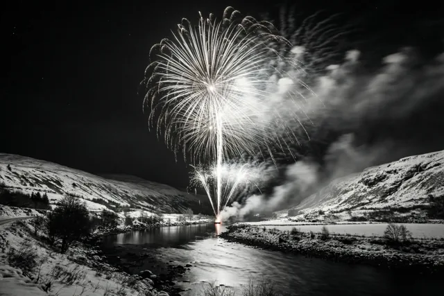 Op deze zwart-witfoto barst vuurwerk los boven een rivier die 's nachts door besneeuwde heuvels kronkelt.