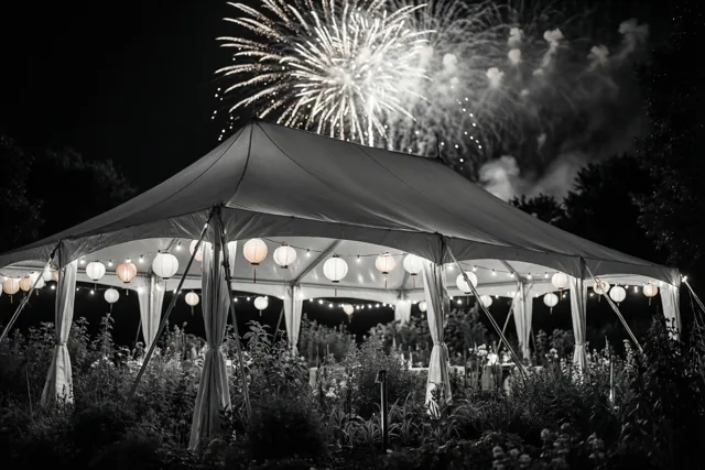 In een nachtelijke tuin met planten op de voorgrond staat een feesttent met lichtsnoeren en papieren lantaarns, terwijl vuurwerk boven het tentdak uitbarst.
