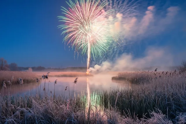 Vuurwerk barst open boven een moeras en weerspiegelt in het water, met berijpt riet op de voorgrond en lage mist onder een diepblauwe hemel.