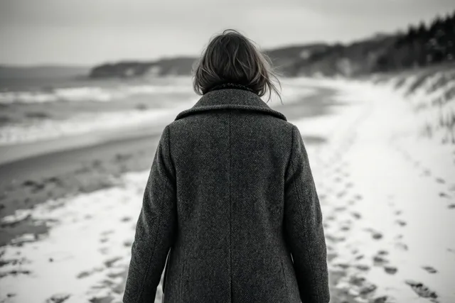 Een persoon in een dikke jas loopt met de rug naar de kijker over een besneeuwd strand naar zee, weergegeven in zwart-wit.