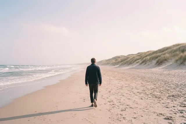 Een persoon loopt met de rug naar de kijker over een zandstrand richting de duinen, met links golven en daarboven een bleke hemel.