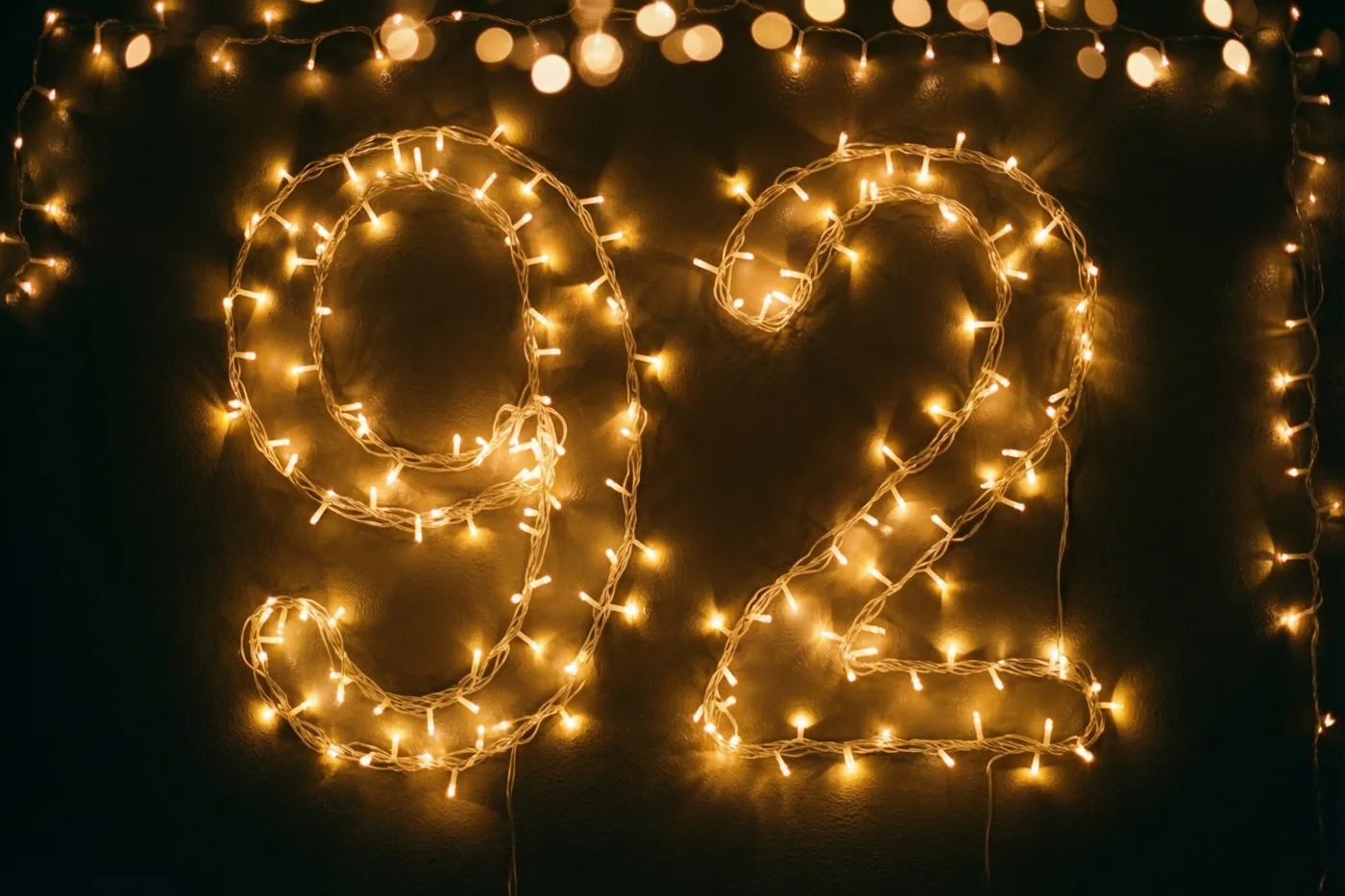 Warmgele lichtjes vormen 92 tegen een zwarte muur, met zachte onscherpe lichtpunten op de achtergrond.