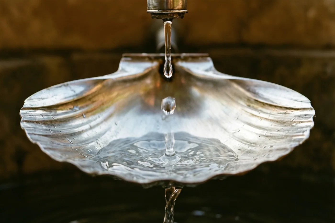 Een enkele waterdruppel valt uit een kraan in een zilveren schelpvormige schaal, met een onscherpe achtergrond in warme bruine tinten.