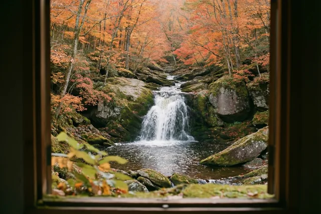 Door een raam is een kleine waterval te zien die over bemoste grijze rotsen stroomt, tussen oranje en rode bladeren in een herfstbos.