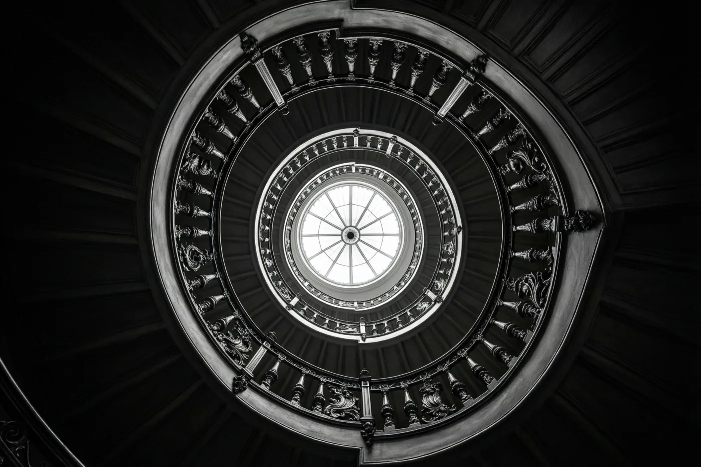 Een zwart-witfoto toont van onderaf een wenteltrap met donkere, sierlijke balustrades en treden rond een rond dakraam.