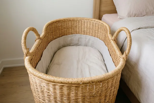 Een leeg rieten wiegmandje met beige linnen staat op een houten vloer naast een bed met bijpassend beddengoed in zacht licht.