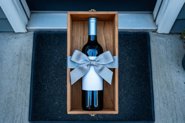 Een fles wijn met een lichtblauwe strik staat in een houten kistje op een donkere deurmat bij een drempel.
