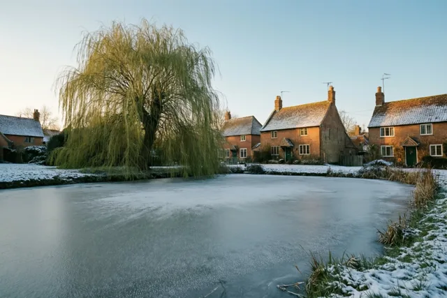 Een bevroren dorpsvijver ligt in de sneeuw, met links een grote wilg en rechts traditionele bakstenen huizen in zacht winterlicht.