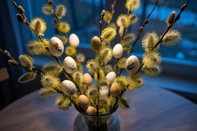 Een vaas met wilgentakken, zachte gele katjes en versierde paaseieren staat op een tafel voor een blauw raam.