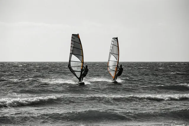 Twee windsurfers varen in de verte naast elkaar over een woelige zee op een zwart-witfoto.