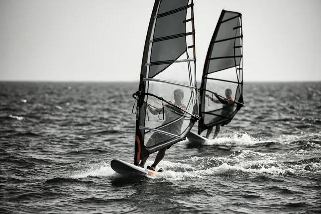 Twee windsurfers varen in de verte naast elkaar over een woelige zee, op een zwart-witbeeld met subtiele oranje accenten op de planken en zeilen.
