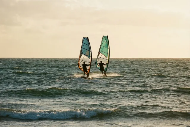 Twee windsurfers varen in de verte naast elkaar over een woelige zee onder een bewolkte hemel, met gedempte blauwe en groene tinten in het water en de zeilen.