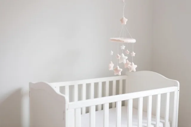 Een wit houten babybedje staat in een sober ingerichte babykamer in wit en zachtroze, met een zachte stervormige mobiel erboven.