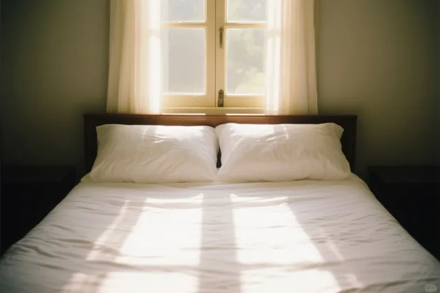 Een wit bed met twee kussens staat onder een raam met doorschijnende gordijnen in een kamer met grijsgroene muren en donker houten meubels, terwijl zonlicht over de sprei valt.