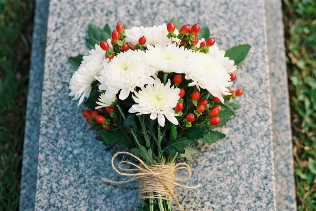 Een met raffia gebonden boeket witte chrysanten en rode bessen ligt op een granieten graf met groen gras langs de randen.