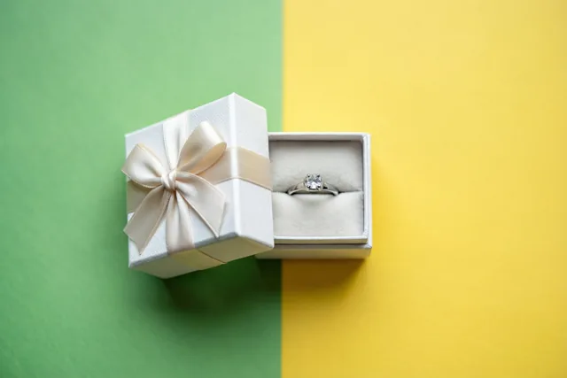 Een open wit geschenkdoosje met een crèmekleurige satijnen strik toont een zilveren ring met een centrale diamant op een zachte voering, tegen een achtergrond met een groen en een geel vlak.