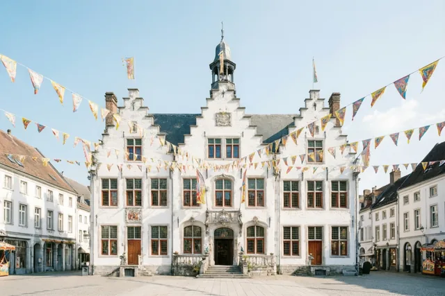 Een wit stadhuis met trapgevels, rode raamkozijnen en een centrale ingang staat aan een bestraat plein met carnavalsvlaggen onder een helderblauwe hemel.