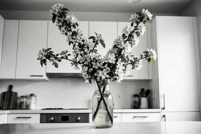 Een hoge glazen kan met een tak witte appelbloesems staat op een wit keukenwerkblad, met witte kasten en een neutrale achtergrond.
