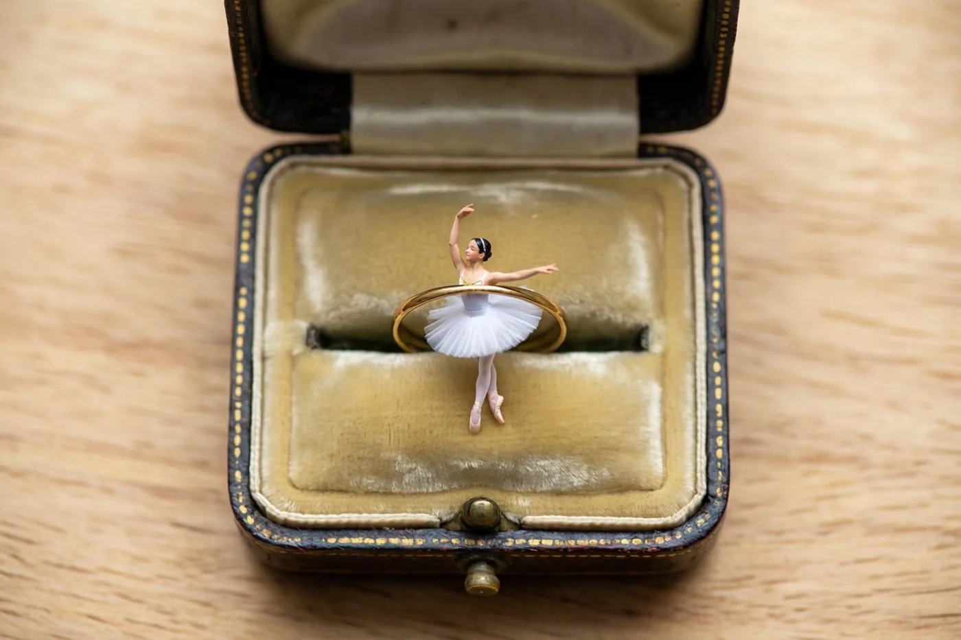 Een miniatuurballerina in een witte tutu staat sierlijk in een open vintage ringdoosje op een houten ondergrond, in zacht licht met warme beige en bruine tinten.