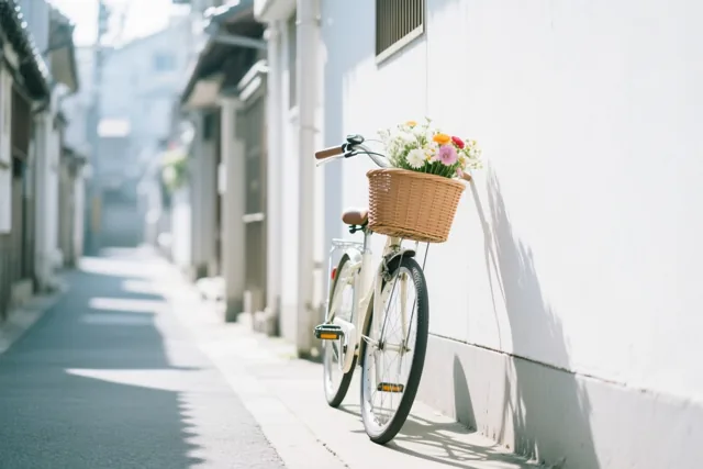 Een witte fiets met een rieten mand vol kleurrijke bloemen leunt tegen een witte muur in een smal, zonnig steegje.