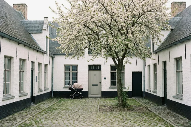 Een kinderwagen staat bij een deur op een binnenplein met kasseien, witte begijnhofhuisjes met donkere leien daken en een boom met witte bloesems.