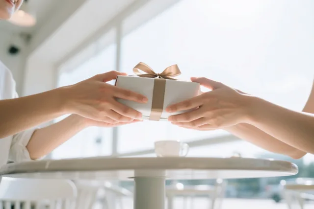 Twee handen geven een witte cadeaudoos met een beige lint door boven een cafétafel tegen een lichte achtergrond.