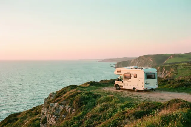 Een witte camper staat bij zonsondergang op een grasrijke klif boven de zee, met roze en blauwgroene pasteltinten in lucht en water.