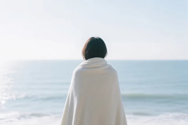 Een persoon met kort donker haar staat met de rug naar de kijker in een witte deken op het strand en kijkt naar een kalme zee onder een heldere hemel.
