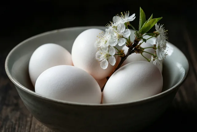 Een keramieken kom met witte eieren en een takje witte bloesems met groene blaadjes staat tegen een donkere houten achtergrond.