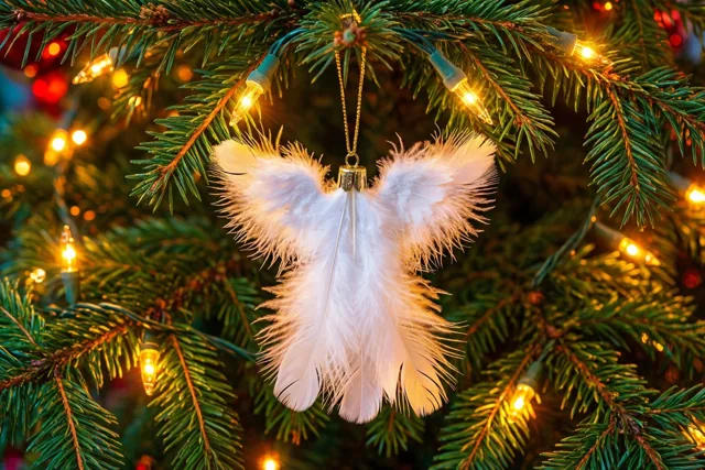 Een wit engelenornament van veren hangt aan een dennentak tussen warm gloeiende kerstlichtjes, met groen, wit en goudgeel als overheersende kleuren.