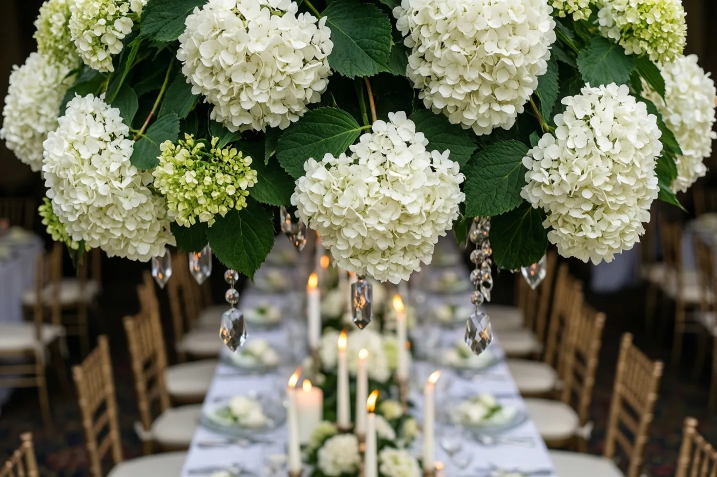 Een lange trouwtafel met brandende kaarsen en kristallen versieringen staat onder een weelderige wolk hangende witte hortensia’s, in witte en groene tinten.