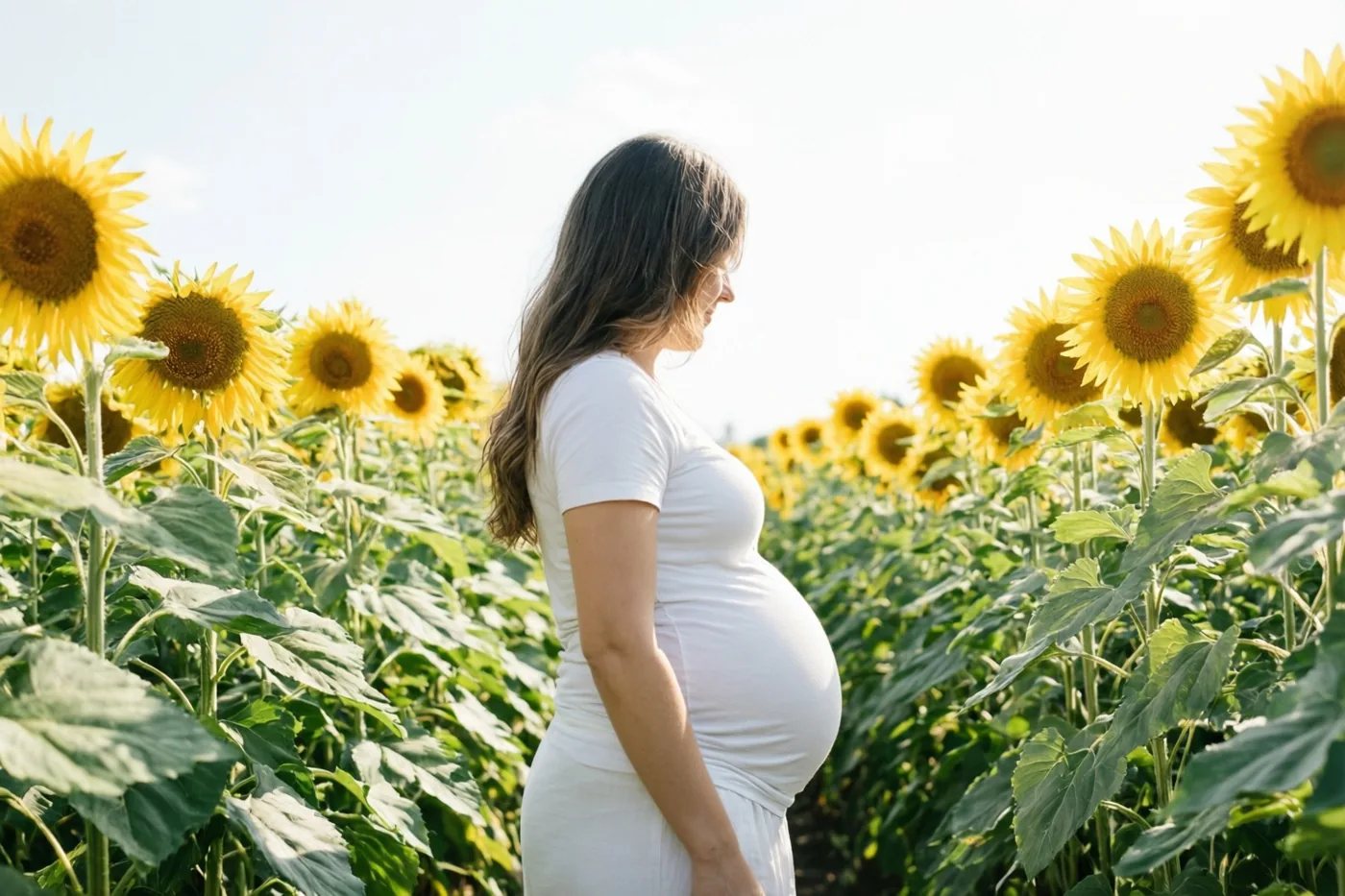 Een zwangere vrouw in een witte jurk wandelt van opzij gezien door een veld met hoge gele zonnebloemen en groen gebladerte onder een heldere hemel.