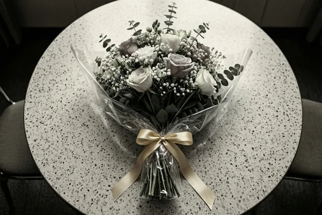 Een boeket witte en mauve rozen met gipskruid en eucalyptus, verpakt in doorzichtig cellofaan met een crèmekleurig lint, staat op een gespikkelde formicatafel in gedempte tinten.